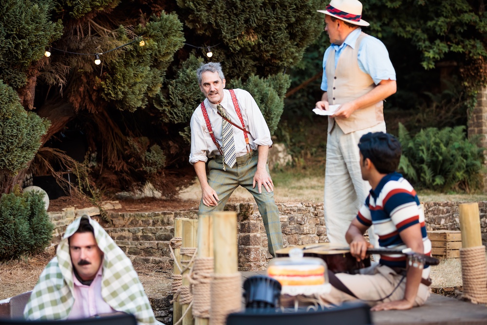 Owen Oakeshott as the Prince of Navarre, standing among fellow cast members in the dry, tree-lined open-air setting of Love’s Labour’s Lost.